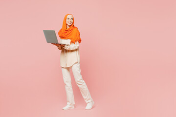 Full body side view young arabian asian muslim IT woman wearing orange abaya hijab holding use work on laptop pc computer isolated on plain pink background. Uae middle eastern islam religious concept.