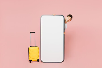 Full body traveler man hold yellow suitcase big huge blank screen mobile cell phone isolated on plain pink background. Tourist travel abroad in free time rest getaway. Air flight trip journey concept.