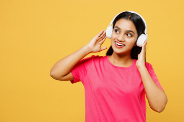 Young minded smiling fun happy Indian woman wearing pink t-shirt casual clothes listen to music in headphones look aside on area isolated on plain yellow background studio portrait. Lifestyle concept.