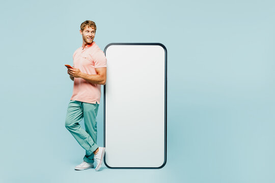 Full Body Sideways Young Man Wear Pink T-shirt Casual Clothes Looking At Big Huge Blank Screen Mobile Cell Phone With Workspace Area Use Smartphone Isolated On Plain Blue Background Lifestyle Concept