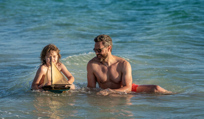 Fototapeta premium summer vacation of friendly family dream. Son and father playing in summer sea. Father and son on sea. Exploring sea with a toy ship as father and son. Summer family fun