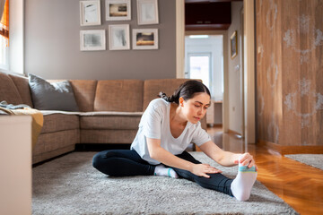 Young Caucasian woman wearing sports clothes sits on floor in bedroom and does stretching bends on...