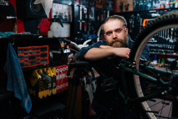 Medium shot portrait of handsome cycling repairman with beard standing leaning on bicycle in repair bike workshop with dark interior, looking at camera. Concept of professional maintenance of bicycle.
