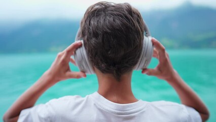 Back of young boy putting headphones while standing at scenic lake shire view. Child listening to music, audiobook, or podcast staring at nature - Powered by Adobe