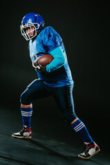 Portrait of a man in a blue uniform for american football runs with a ball on a black background. 