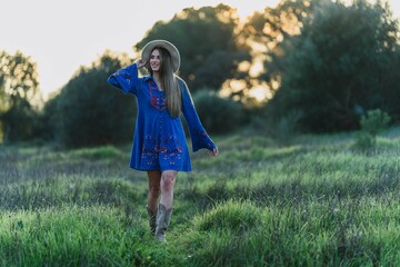 Pretty Caucasian young woman in a blue dress and a hat standing in a sun-kissed meadow