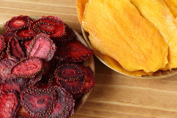 Dried strawberry pieces and dried mango pieces lie on wooden saucers that stand on a wooden table. View from above