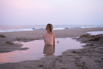 Back view portrait of little lonely sad girl sitting in puddle looking at colourful sky, beautiful...