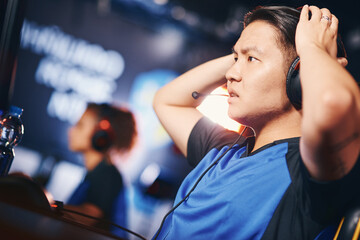 Side view of a confused asian guy, male cyber sport gamer wearing headphones looking at PC screen...