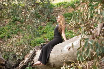 Blonde, young and beautiful woman dressed in black and sitting on the trunk of a fallen eucalyptus tree in the forest, the woman is relaxed and resting on the tree. Concept relaxation and nature.
