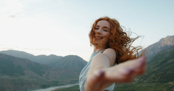 Gorgeous caucasian girl wearing a light dress is posing for a photo shoot on scenic mountain background while wind is blowing her red hair. Follow me and hold my hand. Copy Space
