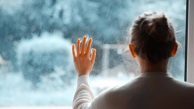 Young caucasian woman touches hands to the window glass in winter, snowing outside .