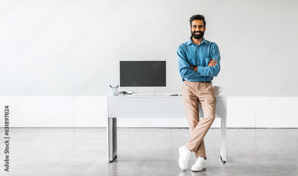 Poster confident indian businessman sitting on desk with folded arms showing computer with blank screen in 