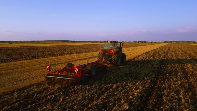 Aerial view of a Contemporary Red Combine tractor plowing and tilling field during sunset. City outskirts farming fimed from a bird's eye view. Tracking shot. 
