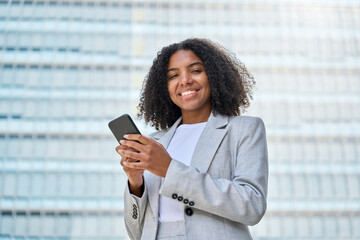 Young happy professional African American business woman wearing suit holding cell phone, looking at cellphone, using mobile apps on smartphone standing on city street, portrait.