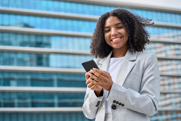 Young happy professional African American business woman wearing suit holding cell phone, looking at camera with cellphone, using mobile apps on smartphone standing on city street, portrait.