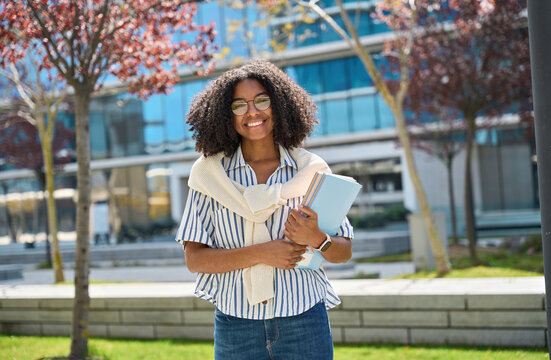 Happy pretty female African American student walking outside university. Smiling cheerful girl model with curly hair wearing glasses, holding notebooks looking at camera outdoors, portrait.