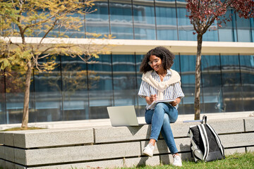 Happy African American girl university or college student elearning online outdoors using laptop...