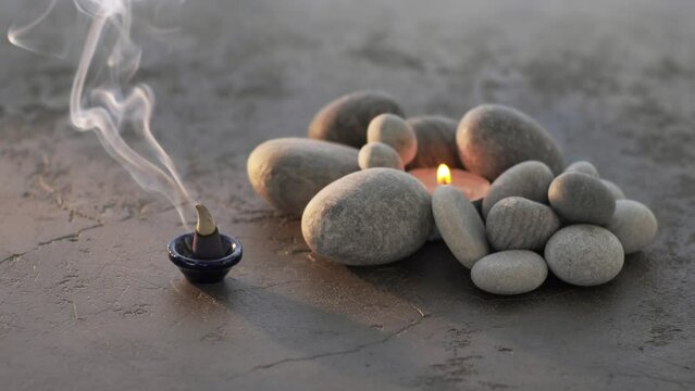 Burning incense cone with aromatic smoke and pebble stones with tea-candle on background