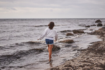Fototapeta na wymiar Teenage girl walking alone along water line on the sand and stone beach of North sea on the coast of Denmark during her summer vacation in scandinavian countries and enjoying water, wind and fresh air