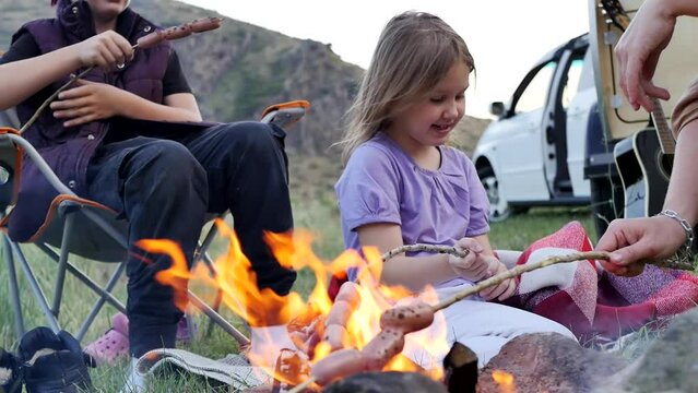 Children and mother fry meat sausages on an open fire with the help of shrub branches. Family picnic in the mountains on a road trip by car with trailer camper. Vanlife and active lifestyle