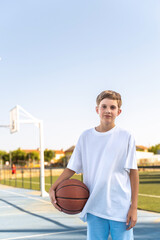 Young boy posing with a basketball ball while standing outdoors at sports court. Sports concept.