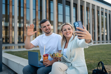Smilling joyous colleagues taking selfie and waving while eating lunch outside office building.