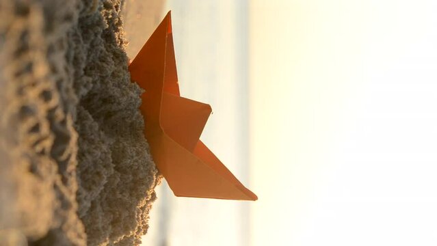 Paper orange boat on sand near sea waves with sunny path on sandy beach of sea shore during sunset dawn with shining sun close-up. Swimming dream sailing traveling vacation tourism concept Vertical