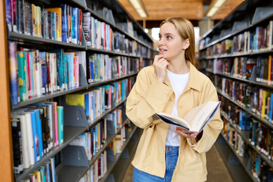 Smart pretty focused blonde girl student holding book looking away standing in modern university campus library or bookstore thinking of college course study reading literature for doing research.