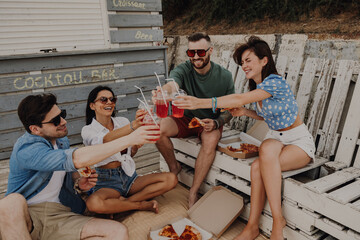 Happy young friends toasting with cocktails while enjoying pizza outdoors together
