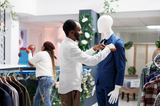 African american man dressing mannequing in formal male outfit in clothing store. Fashion boutique employee adjusting luxury suit on dummy model in department shopping mall