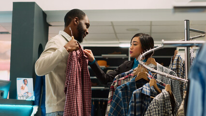 Store assistant giving clothes to man to try on in clothing boutique, helping person to buy trendy shirts. Employee talking to customer about new modern collection, shopping center. Handheld shot.
