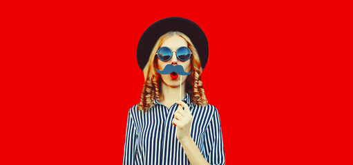 Portrait of funny cheerful young woman having fun showing mustache on stick on red background