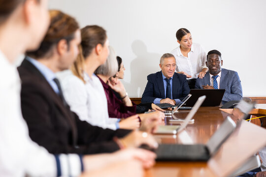 Multiracial group of friendly mature male senior executives in formal suits holding business team meeting in conference hall at office while young white female company secretary assisting them