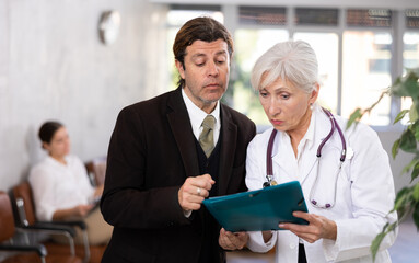 Focused adult manager of modern clinic discussing documents with experienced senior female doctor while standing in hall with folder in hands