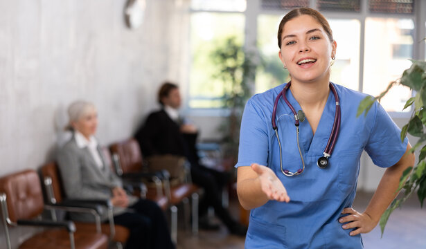 Cheerful friendly young female doctor in blue uniform making welcome gesture, politely inviting patients in medical office