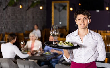 Confident smiling young waiter in maroon apron standing with serving tray, inviting to taste delicious dishes in modern cozy restaurant