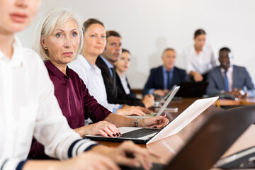 Side view of senior white business woman in burgundy satin blouse sitting among colleagues at table in conference room and interestedly listening to speaker's presentation during corporate group