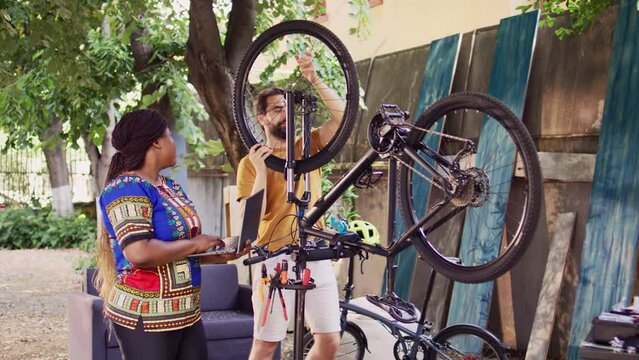 African american woman searching on minicomputer to help young man repair damaged bike. Sports-loving couple fixing bicycle chain while using laptop for annual summer outdoor cycling.