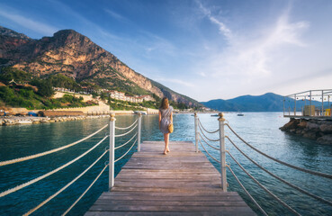 Young woman on wooden pier on sea shore is looking on mountain, blue water and sky at sunset in summer. Girl on jetty, beach, hotels on the hill, green trees, city, rock, boats in Oludeniz, Turkey