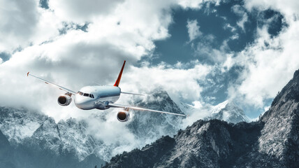 Fototapeta na wymiar Airplane is flying over low clouds against mountains with snowy peaks. Landscape. Passenger airplane, cloudy sky, rocks, snow. Passenger aircraft. Business, commercial travel. Aerial view of plane