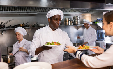 Focused young adult man chef giving out ready meals to waitress on order station in open restaurant kitchen