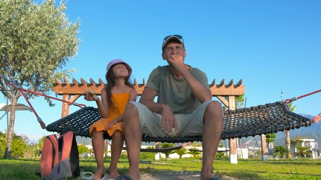 Child and father sitting in hammock. A view of dad with his little daughter enjoy to eat the bun in hammock during summer sea traveling.