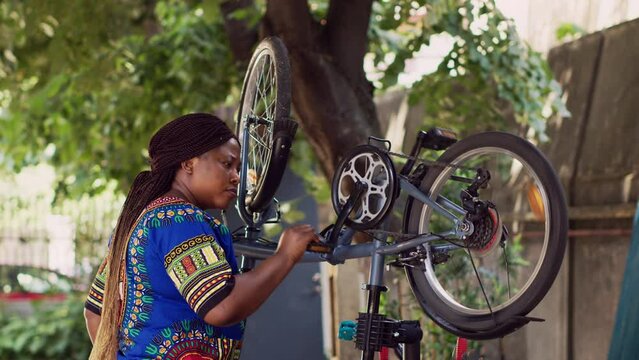 Active african american female cyclist maintaining bike pedal and fine-tunes the gears for outdoor leisure cycling. Motivated healthy black woman holding and reparing modern bicycle in home yard.