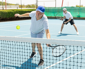 Positive elderly male player serving ball during training tennis in court outdoors