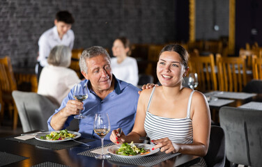 Smiling senior man spending time with young girl in restaurant. Carefree couple laughing merrily, enjoying delicious food and drinking wine at dinner. Age-gap relationships concept