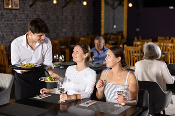 Obraz premium Young male waiter in uniform serves ordered dish to two women in restaurant..