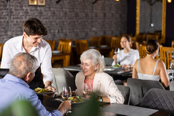 Young guy waiter serves ordered dish elderly couple of man and woman in restaurant