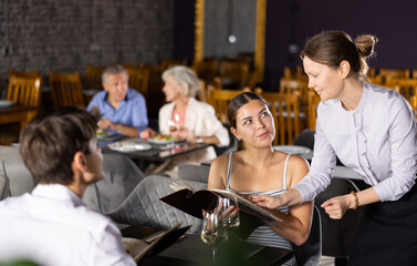 Young male and female colleagues having nice time in sparsely populated cozy restaurant. Waitress answers questions of visitors, helps to choose diet dish for late dinner in menu