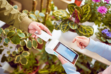 view of hands making contactless payment with card reader between buyer and seller in plant store...